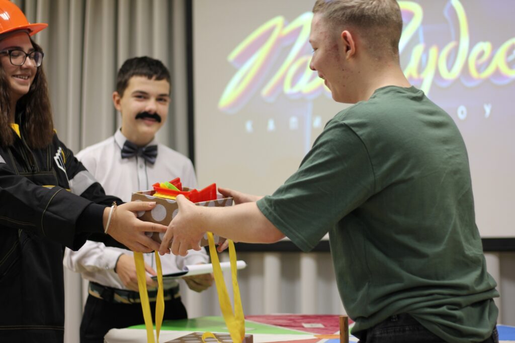 На изображении трое молодых людей. Двое обмениваются призами, третий улыбается в белой рубашке с бабочкой. На заднем плане экран с надписью. На изображении трое молодых людей. Двое обмениваются призами, третий улыбается в белой рубашке с бабочкой. На заднем плане экран с надписью.