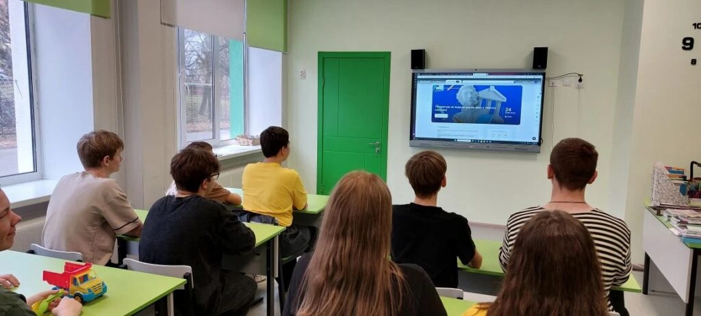 В комнате с большими окнами и зелёной дверью группа людей сидит за партами, смотрит на экран телевизора, на котором что-то показывают. В комнате с большими окнами и зелёной дверью группа людей сидит за партами, смотрит на экран телевизора, на котором что-то показывают.