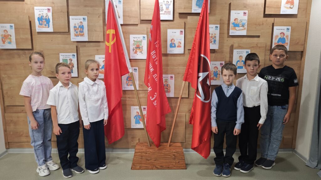 На изображении шесть детей стоят в ряд перед двумя красными флагами. На заднем плане — деревянные панели с детскими рисунками. На изображении шесть детей стоят в ряд перед двумя красными флагами. На заднем плане — деревянные панели с детскими рисунками.