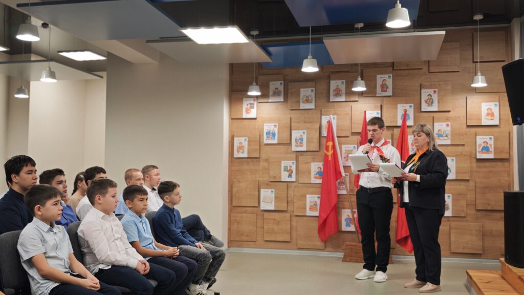 В комнате сидят дети, перед ними стоят двое взрослых с бумагами. На стене висят флаги и фотографии. Помещение освещено подвесными лампами. В комнате сидят дети, перед ними стоят двое взрослых с бумагами. На стене висят флаги и фотографии. Помещение освещено подвесными лампами.