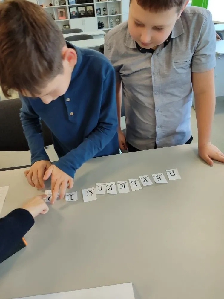 На изображении двое мальчиков стоят у стола. На столе лежат карточки с буквами, образующие слово. Мальчики сосредоточены на карточках. На изображении двое мальчиков стоят у стола. На столе лежат карточки с буквами, образующие слово. Мальчики сосредоточены на карточках.
