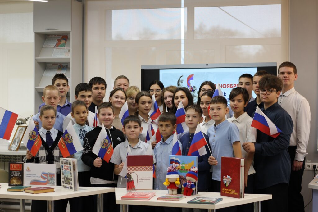 Группа детей с флагами России и книгами на столах. На заднем плане экран с надписью. Помещение светлое, с большими окнами. Группа детей с флагами России и книгами на столах. На заднем плане экран с надписью. Помещение светлое, с большими окнами.