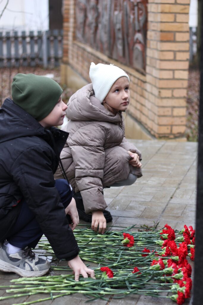 На изображении двое детей в куртках и шапках сидят на корточках. Перед ними лежат красные гвоздики на земле. На заднем плане кирпичная стена. На изображении двое детей в куртках и шапках сидят на корточках. Перед ними лежат красные гвоздики на земле. На заднем плане кирпичная стена.