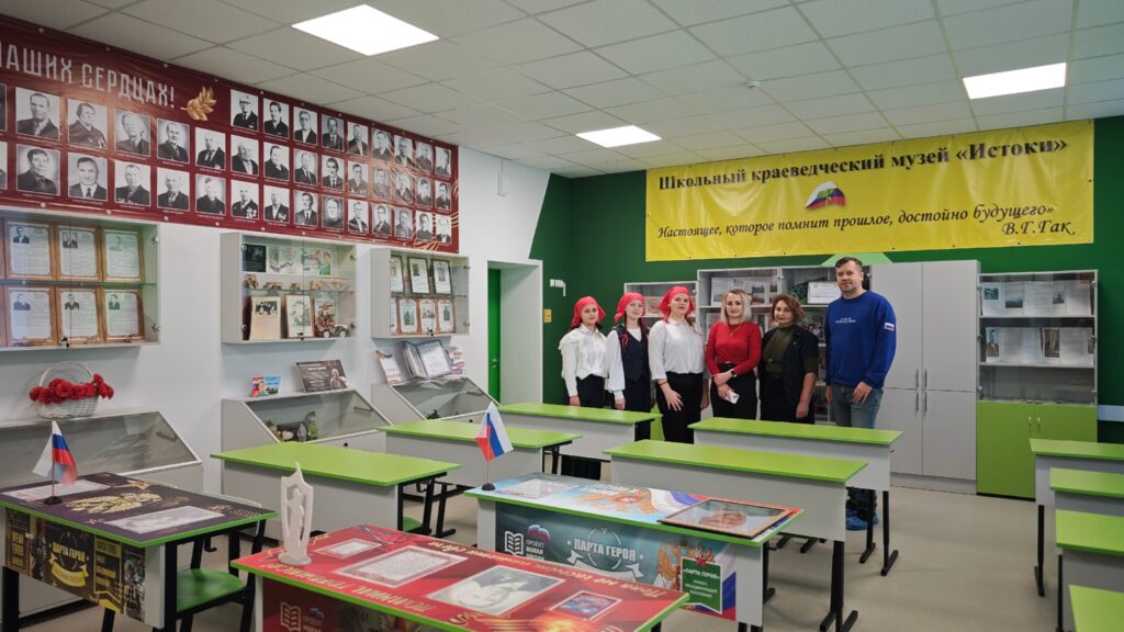 Музей с зелёными столами и стендами, на стенах фотографии и баннеры. Пять человек стоят в центре, одеты в костюмы.