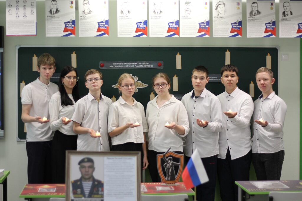 На изображении группа школьников в белых рубашках стоит в классе. На стенах висят плакаты. В центре — портрет и флаг России. На изображении группа школьников в белых рубашках стоит в классе. На стенах висят плакаты. В центре — портрет и флаг России.