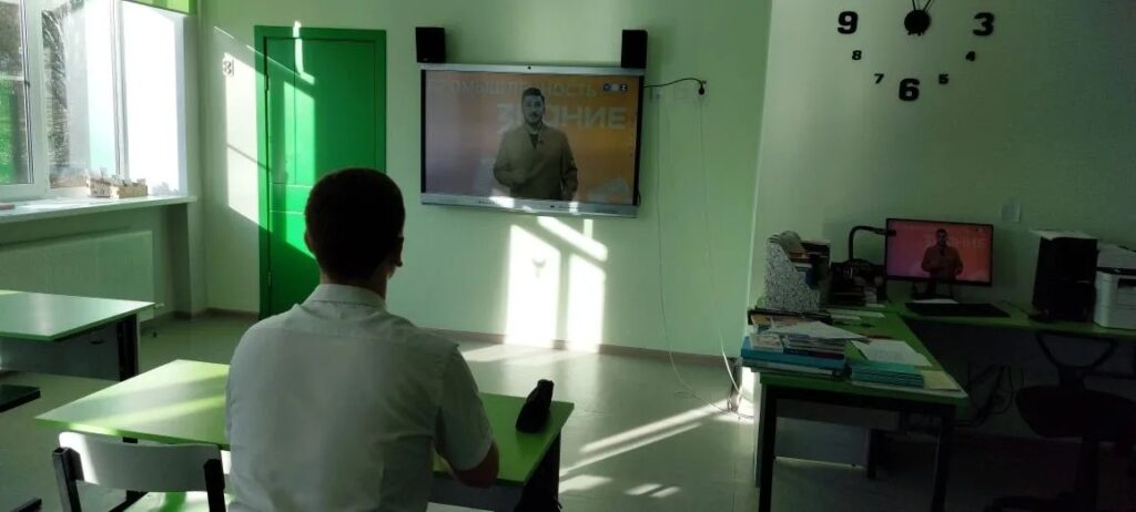 В комнате с белыми стенами и зелёной дверью стоит телевизор, на экране которого человек. На стене часы с цифрами. В комнате есть парты и стулья. На парте лежит ноутбук.