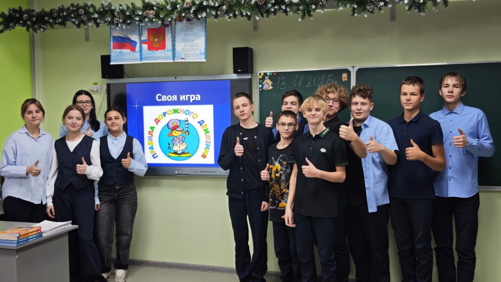 На изображении группа детей в классе. Они стоят перед экраном с надписью «Своя игра» и рисунком. Дети показывают жест «палец вверх».
