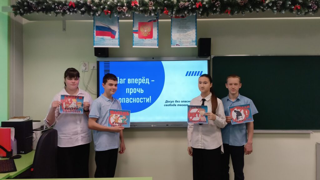 На изображении четыре человека в школьной форме стоят перед экраном. На экране надпись «Вперёд — прочь опасности!». Над экраном висят флаги.