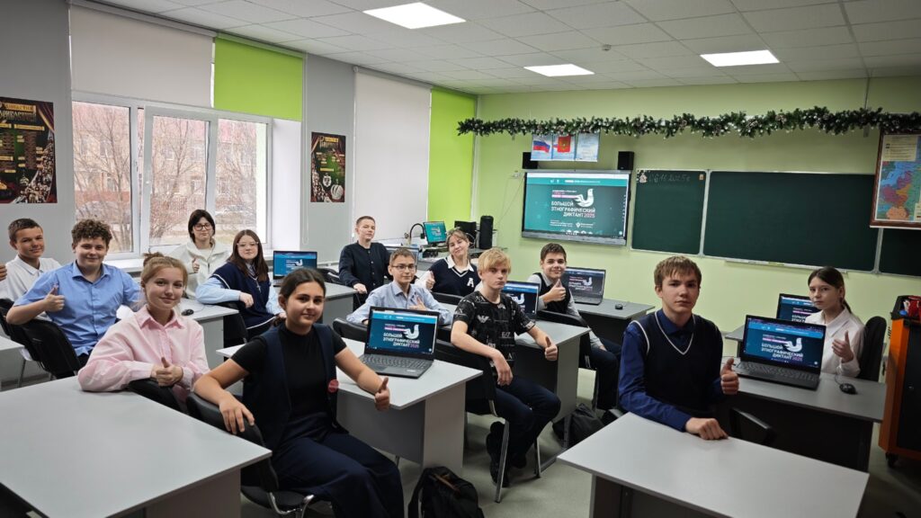 Классная комната с учениками за партами, на которых стоят ноутбуки. На стенах висят плакаты и доска. Светлые стены и окна. Классная комната с учениками за партами, на которых стоят ноутбуки. На стенах висят плакаты и доска. Светлые стены и окна.