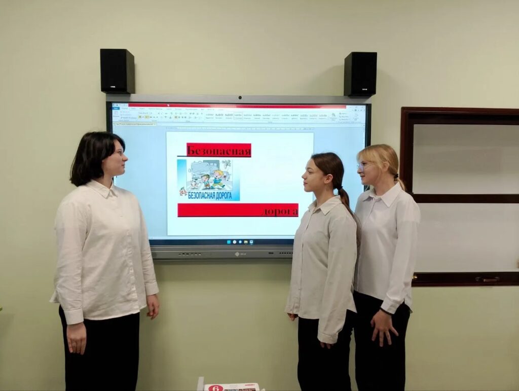 На изображении три человека в белых рубашках стоят перед экраном. На экране текст и картинка. В комнате светло-зелёная стена.