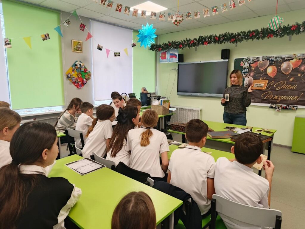 В классе дети в белых рубашках сидят за зелёными партами, слушая женщину у доски. На стенах украшения, на доске плакат.