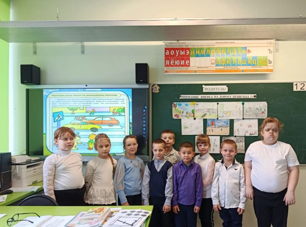 На изображении класс с детьми, стоящими перед доской и экраном. На доске висят рисунки и надписи. В классе зелёная мебель и стены.
