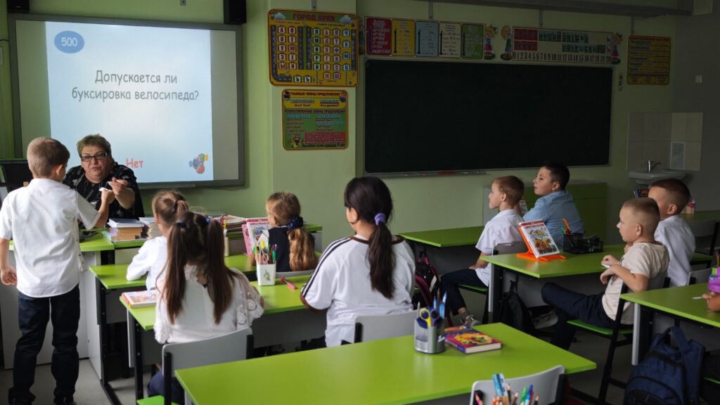В классе дети сидят за партами, слушают учителя. На стене экран с текстом о буксировке велосипеда. Класс украшен яркими плакатами.