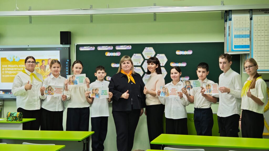 Группа детей и взрослых в классе. Дети держат плакаты. На стенах доска и экран. Классная комната с партами.