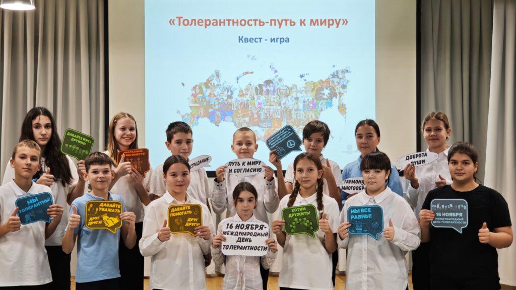 Группа детей стоит в ряд, держа таблички с надписями о толерантности. На заднем плане карта и надпись «Толерантность-путь к миру».