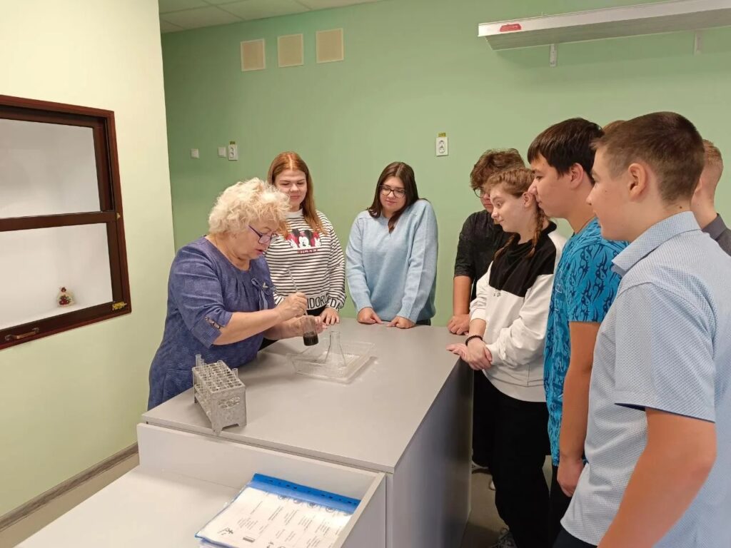 На изображении группа подростков и пожилая женщина в помещении. Женщина что-то показывает в стакане на столе. Стены зелёные. На изображении группа подростков и пожилая женщина в помещении. Женщина что-то показывает в стакане на столе. Стены зелёные.