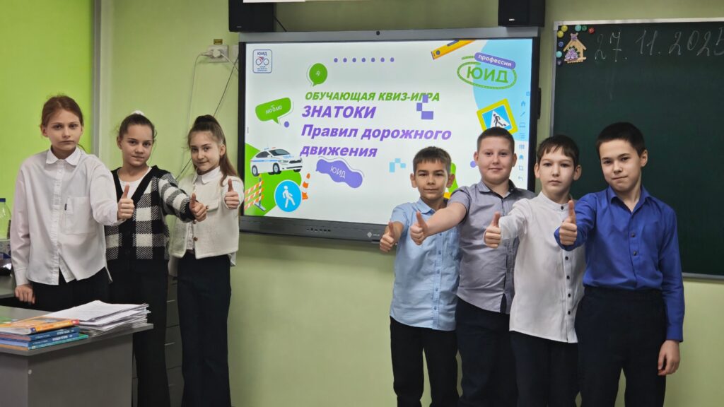 На изображении шесть детей в школьной форме стоят в классе. Они показывают жест «палец вверх». На экране телевизора — игра «Знатоки правил дорожного движения».