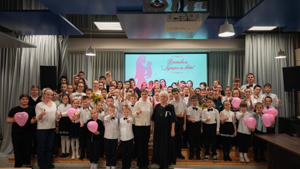 Группа людей стоит в помещении, держа розовые шары. На заднем плане экран с надписью и изображением. Освещение от ламп.