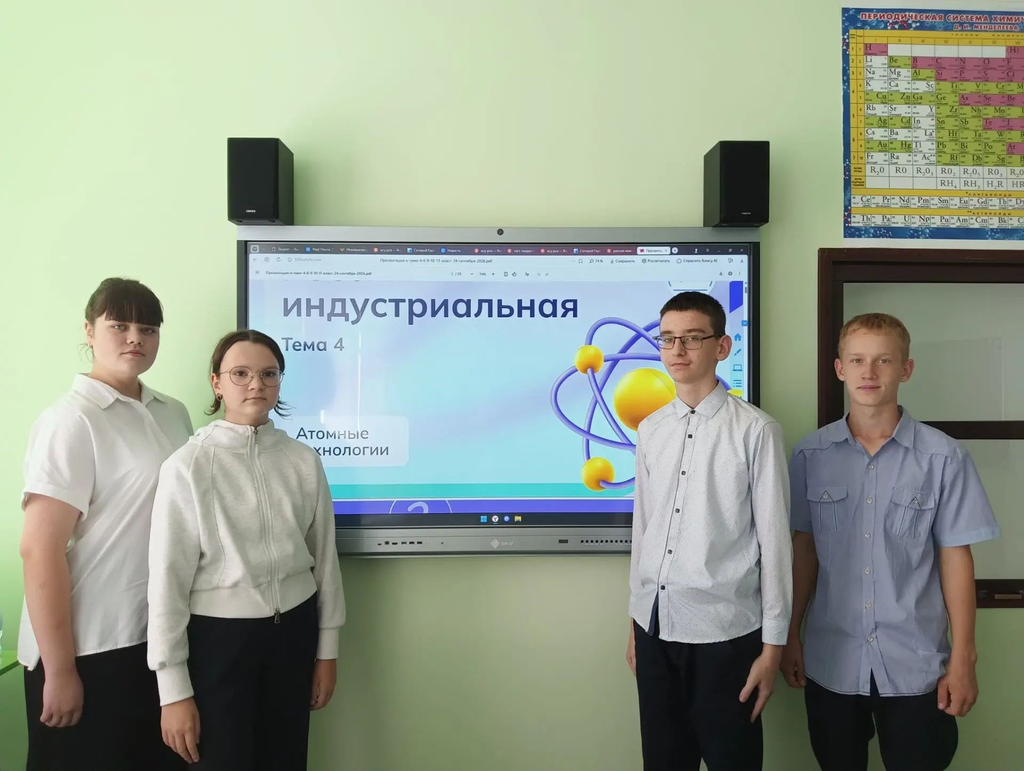На изображении четыре человека в школьной форме стоят перед экраном с надписью «Индустриальная. Тема 4. Атомные технологии». На стене также висит таблица Менделеева.