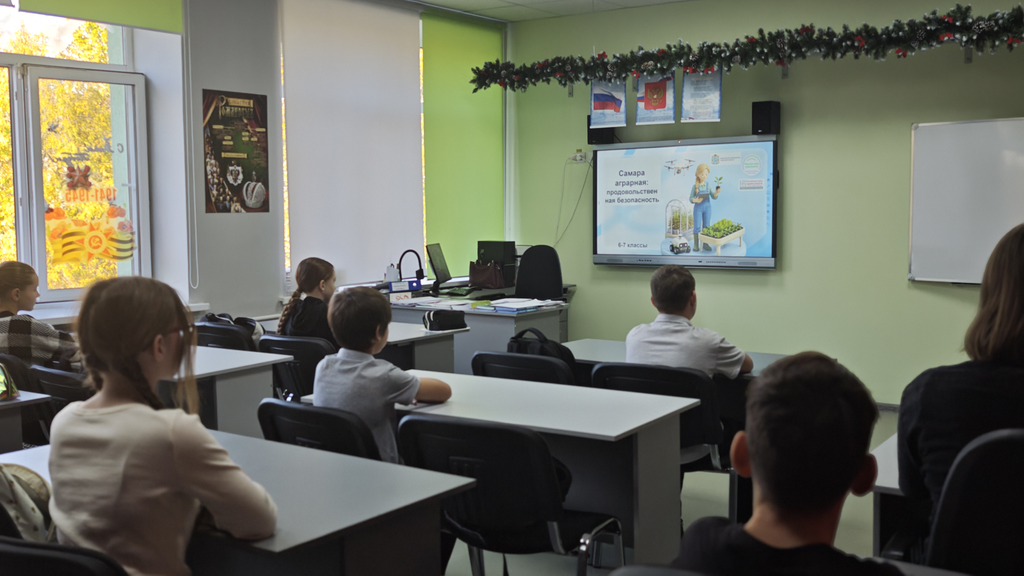 Классная комната с зелёными стенами, украшенная гирляндой. На стене большой экран, на котором презентация. Дети сидят за партами, смотрят на экран.